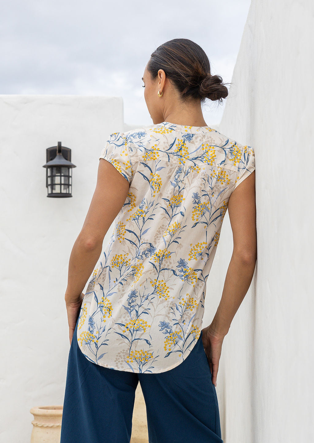 Woman stands backwards wearing a 100% cotton neutral based floral blouse and blue pants.