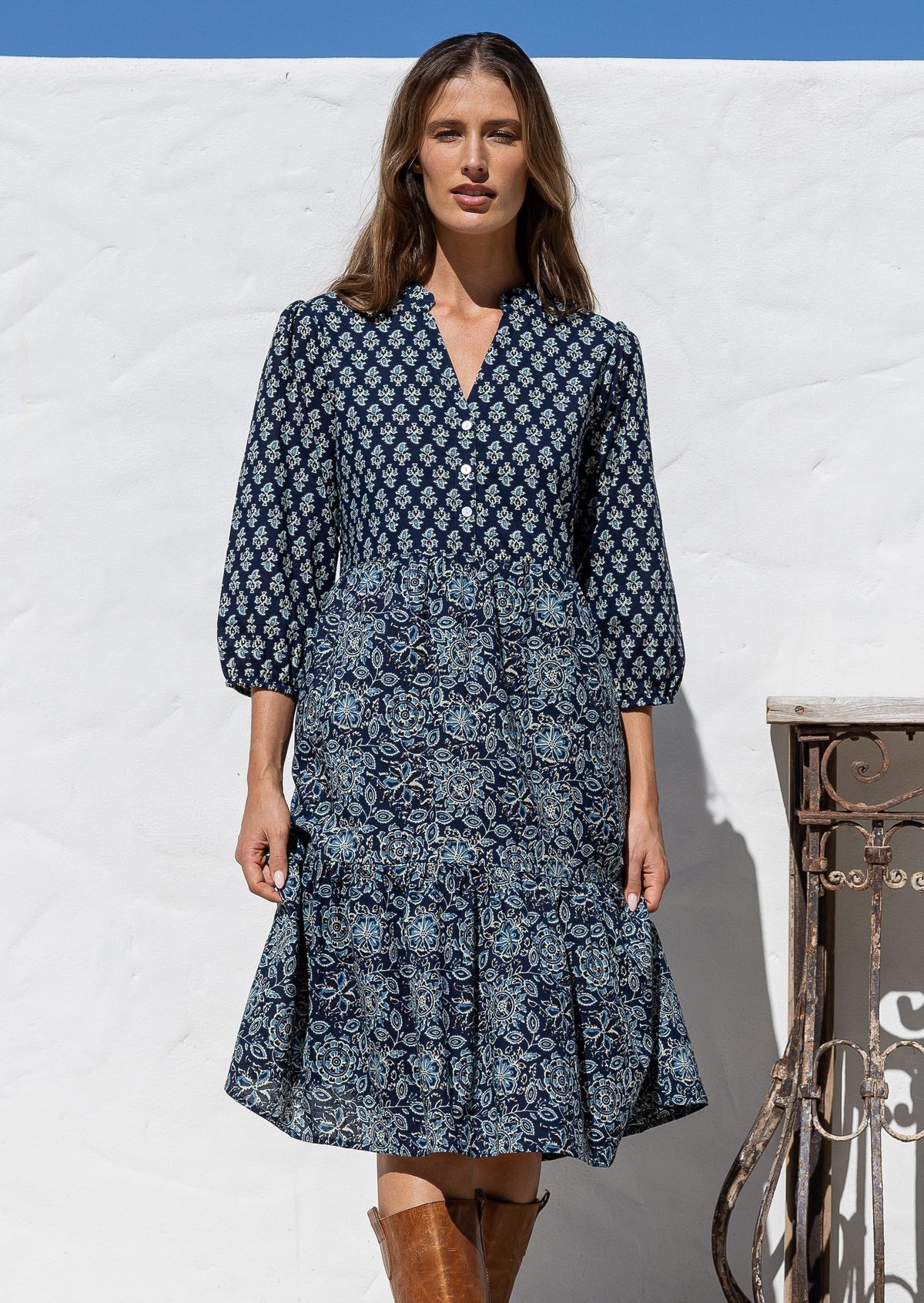 Woman wearing a blue patterned dress and brown boots standing against a white wall.
