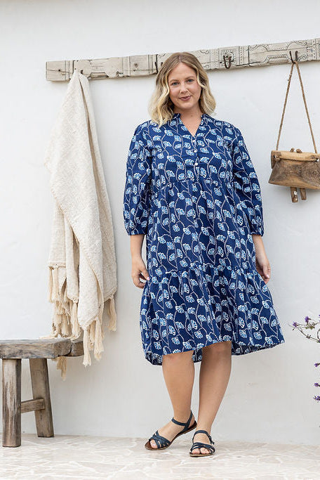 Woman wearing a blue patterned dress and brown boots standing against a white wall.
