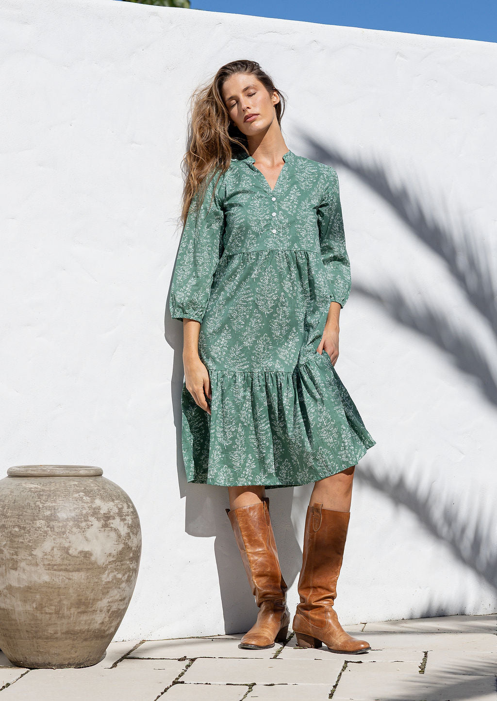Woman in a green floral dress and brown boots standing against a white wall.