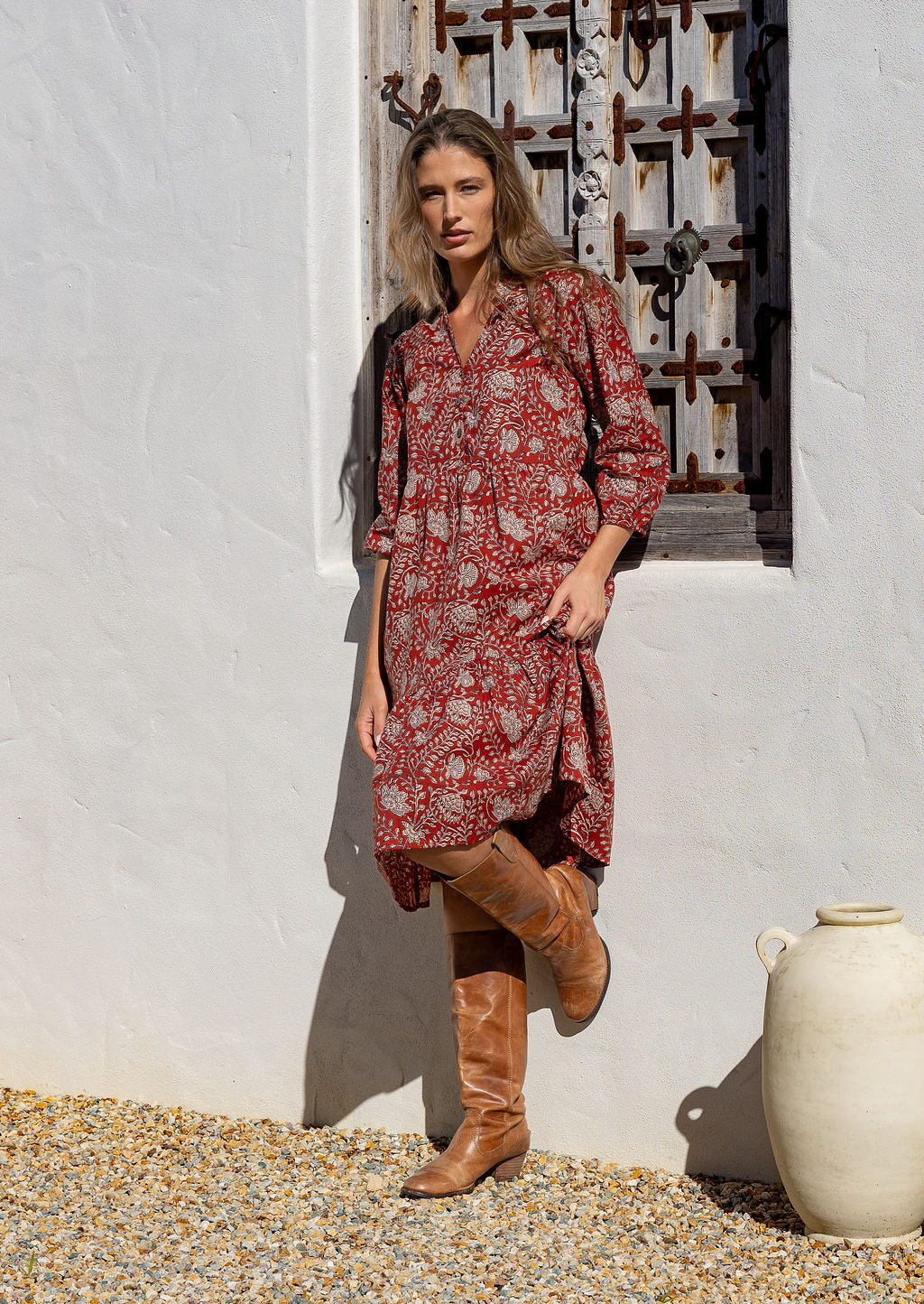 Woman in a red floral dress and brown boots standing against a white wall with a decorative metal gate.
