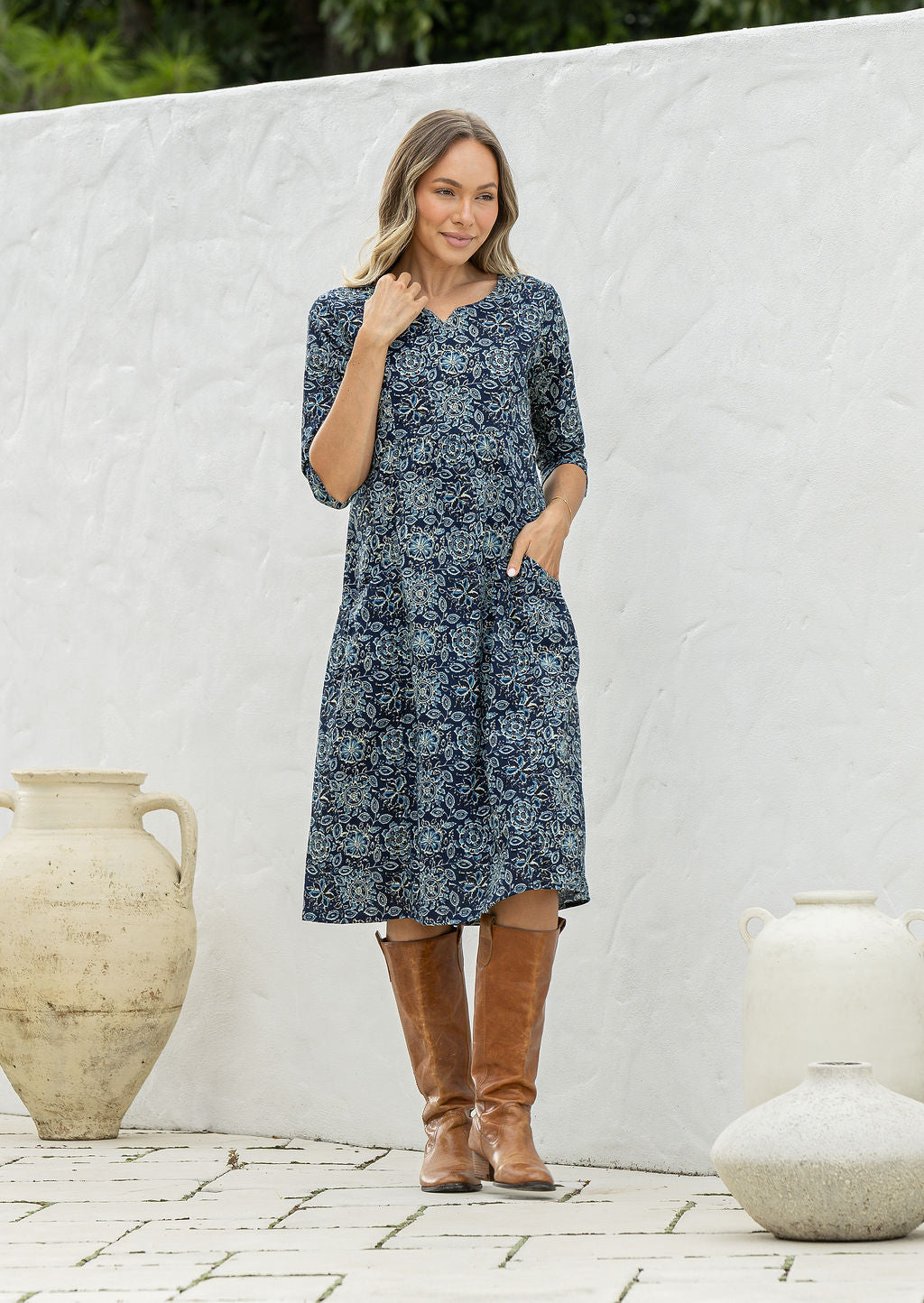 Woman poses in a 100% cotton navy blue based floral dress with sleeves and one hand in pocket
