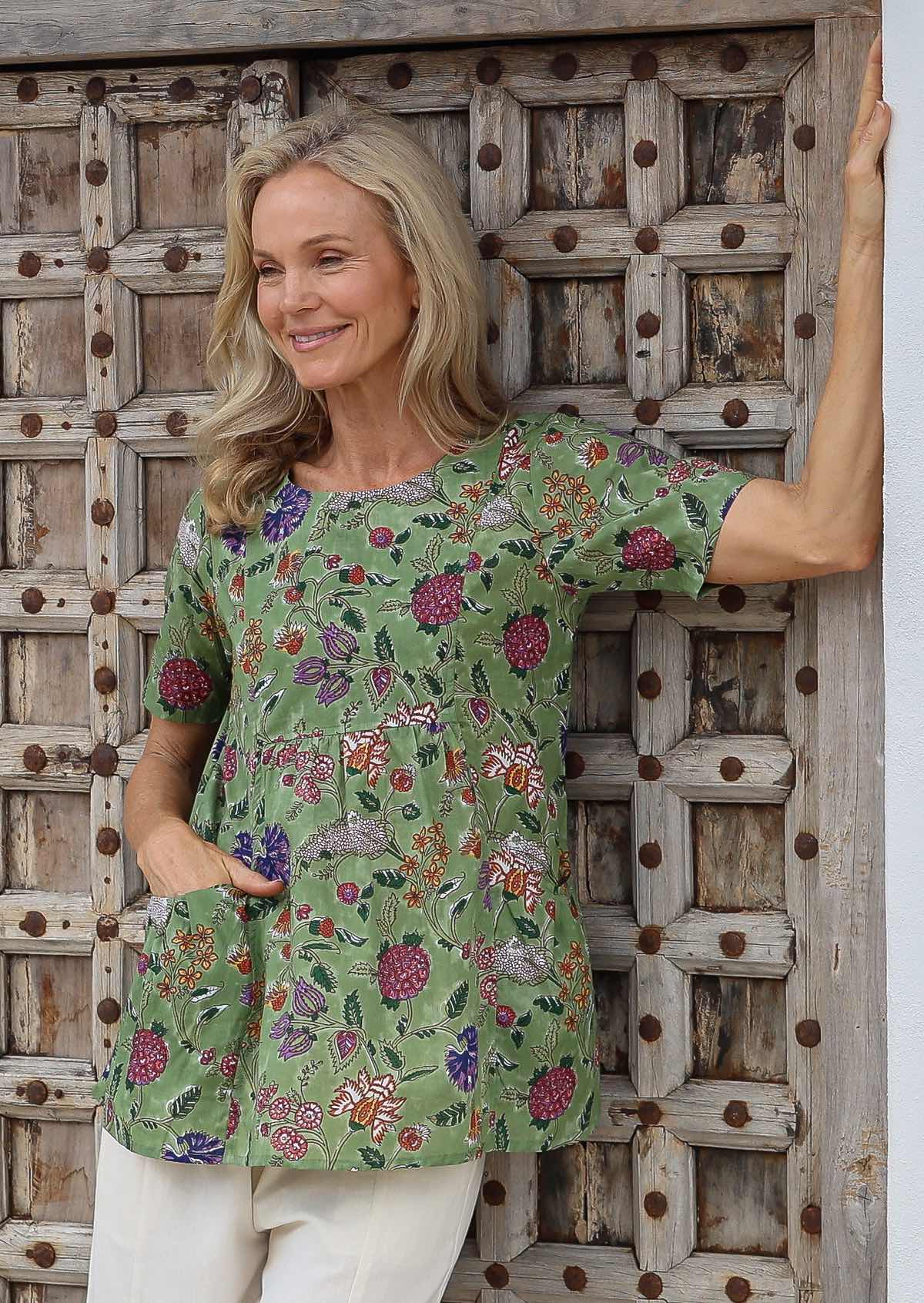 Woman wearing a moss green based multicoloured floral blouse with empire waistline, short sleeves and hand in pocket in front of a wooden door.