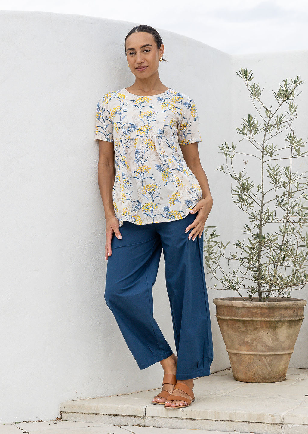 Woman standing wearing a lightweight 100% cotton neutral floral print top with plain blue cotton pants