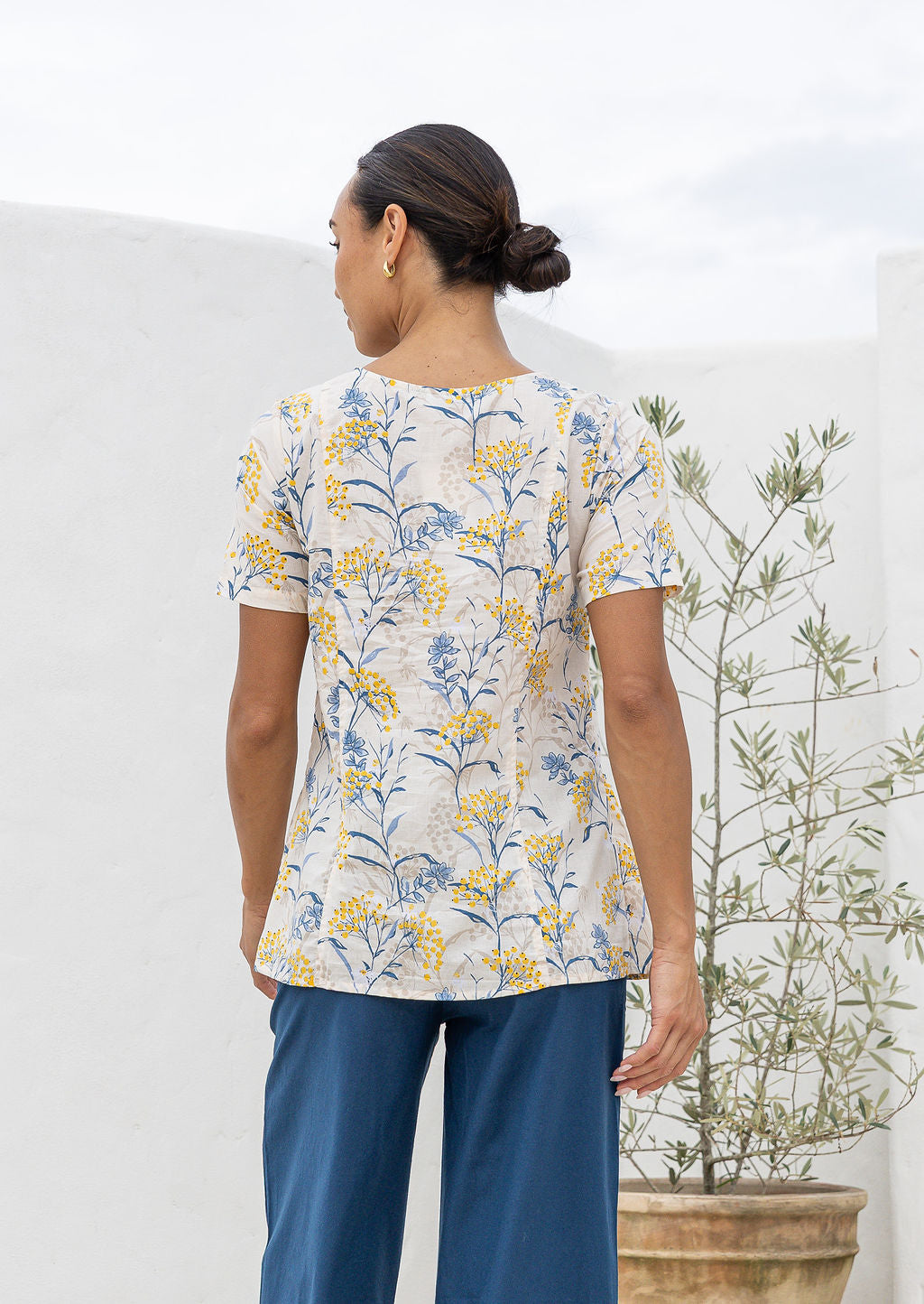 Woman standing backwards wearing a short sleeved neutral based floral cotton top with empire waistline and deep pockets, paired with plain blue cotton pants