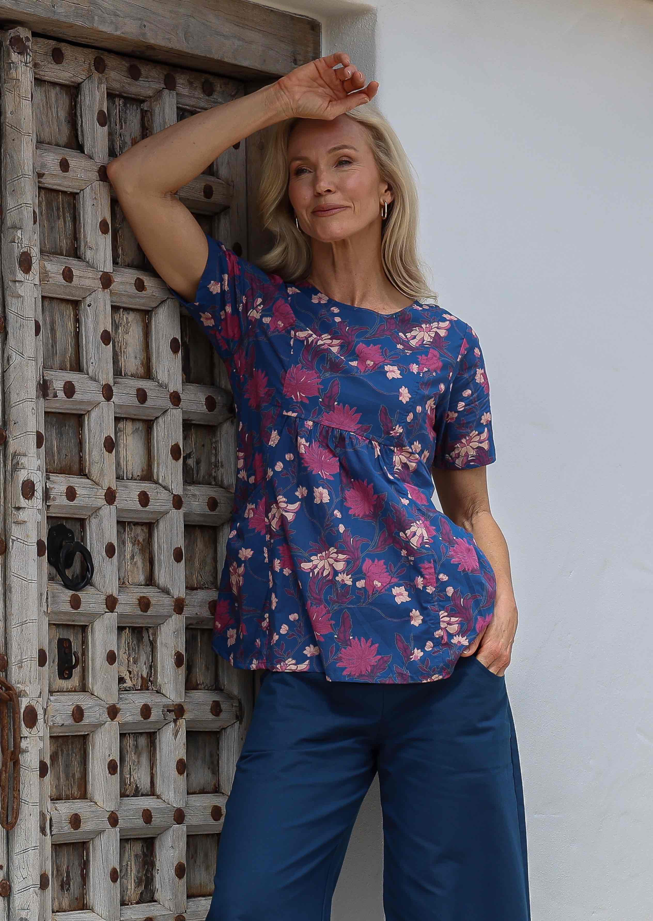 Woman poses in short sleeved blue floral cotton top with empire waistline and deep pockets, paired with sea blue cotton pants