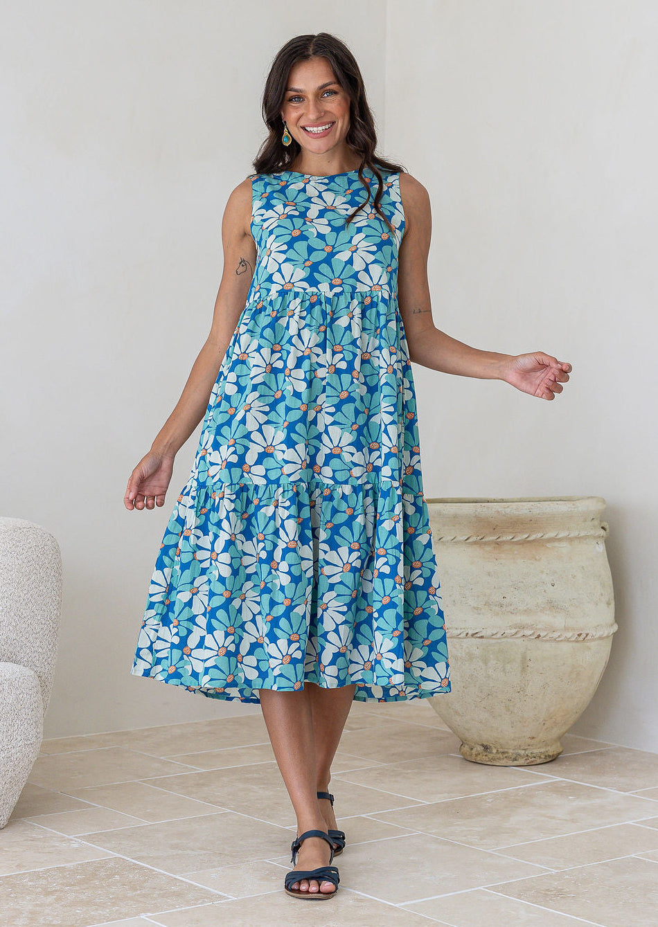 Woman wears cotton sleeveless three tiered midi length dress in aqua and royal blue daisy print
