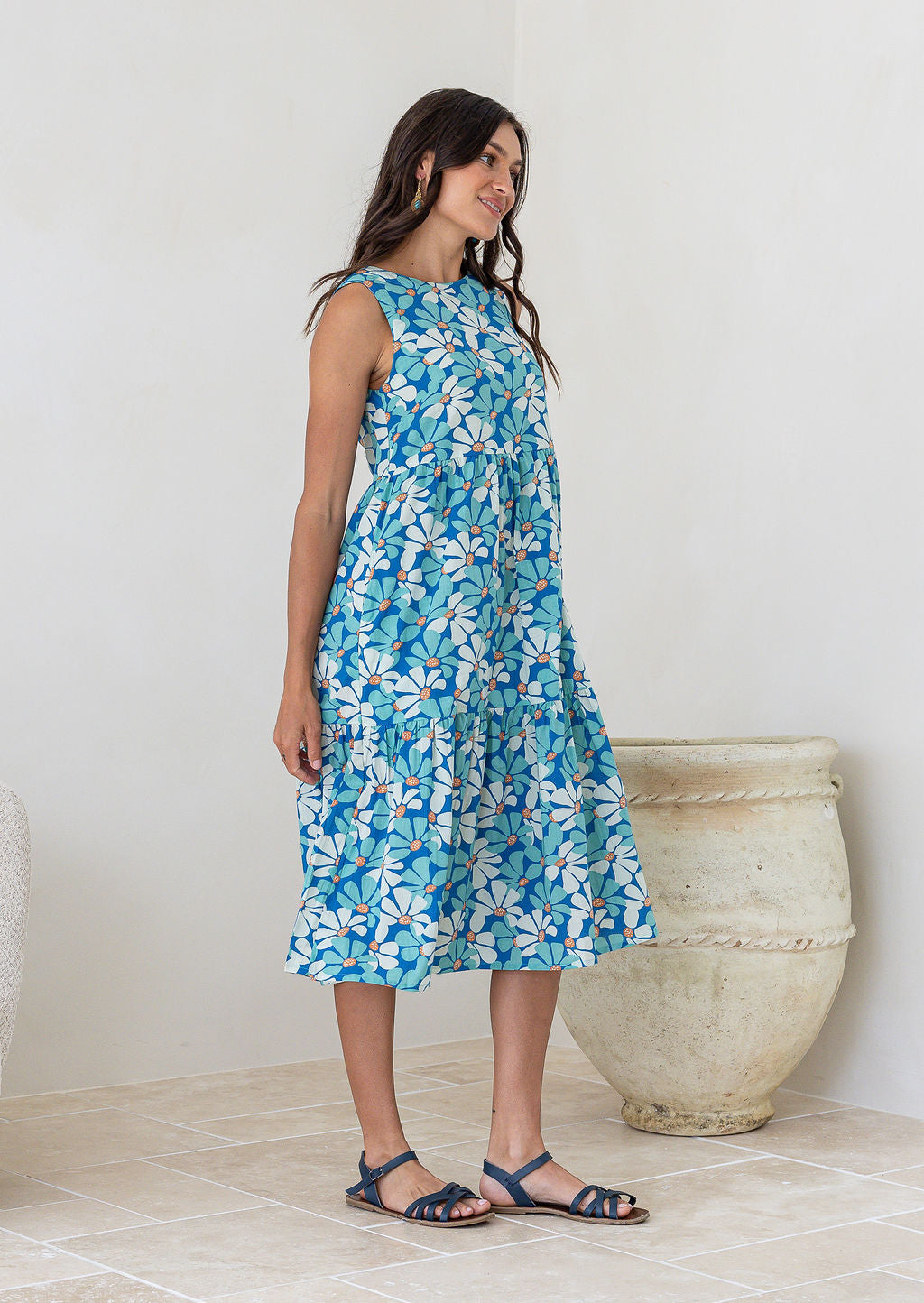 Woman stands side on in cotton sleeveless three tiered midi length dress in aqua and royal blue daisy print