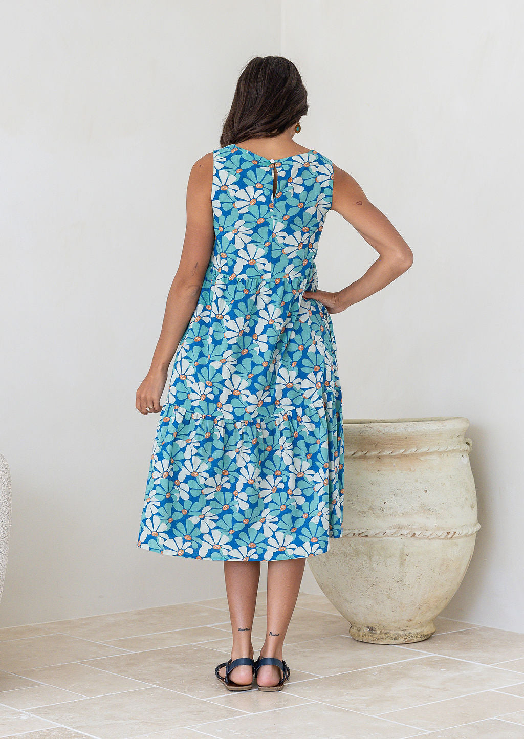 Back view of woman in cotton sleeveless three tiered midi length dress in aqua and royal blue daisy print