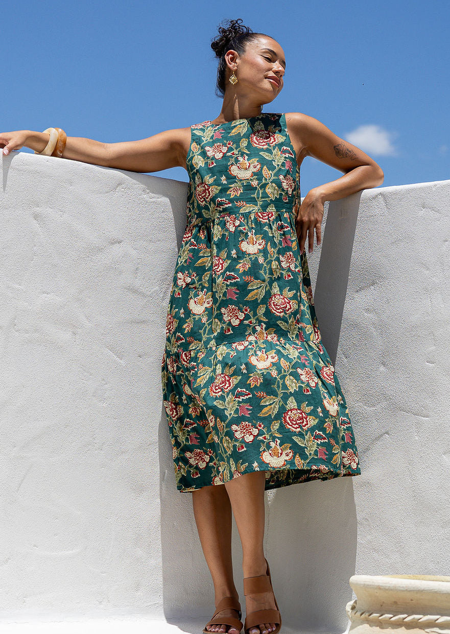 Woman in a sleeveless, floral green based 100% cotton dress standing in front of white wall.