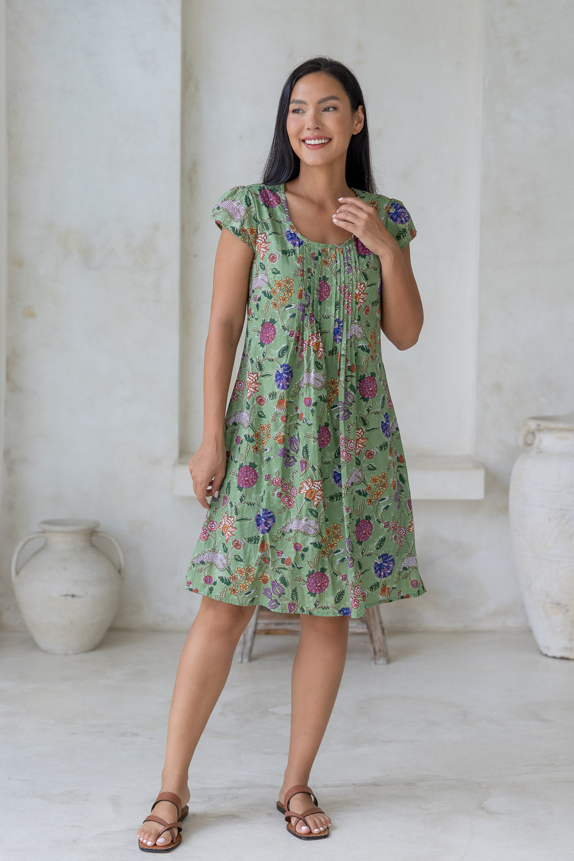 Woman wearing a moss green floral cotton dress with cap sleeves, round neckline and small pleated design across the bodice.