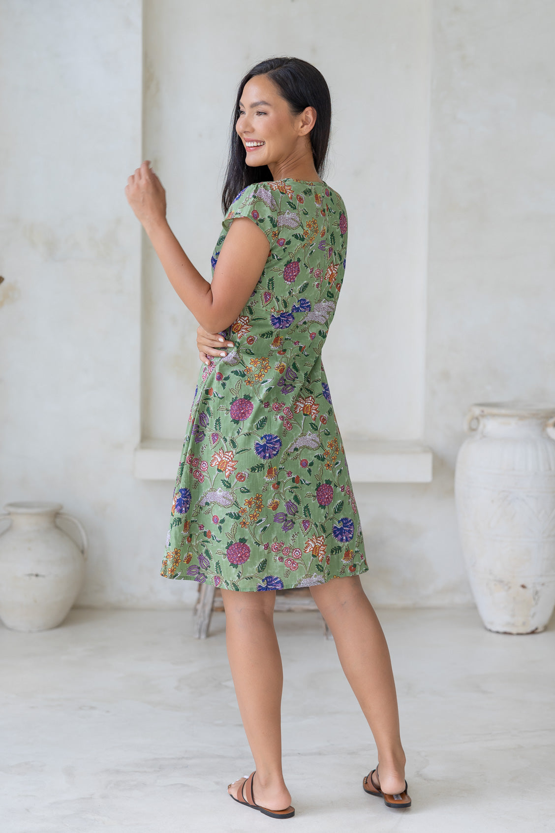 Woman posing in a lightweight 100% cotton above knee dress with cap sleeves, fitted bodice and a-line skirt in moss green folk art style floral print.