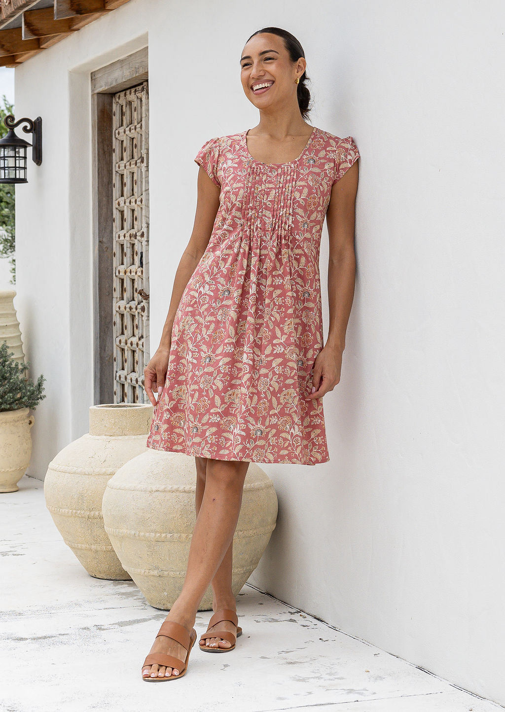 Woman wearing a 100% cotton rose pink based floral dress standing in front of decorative vases.
