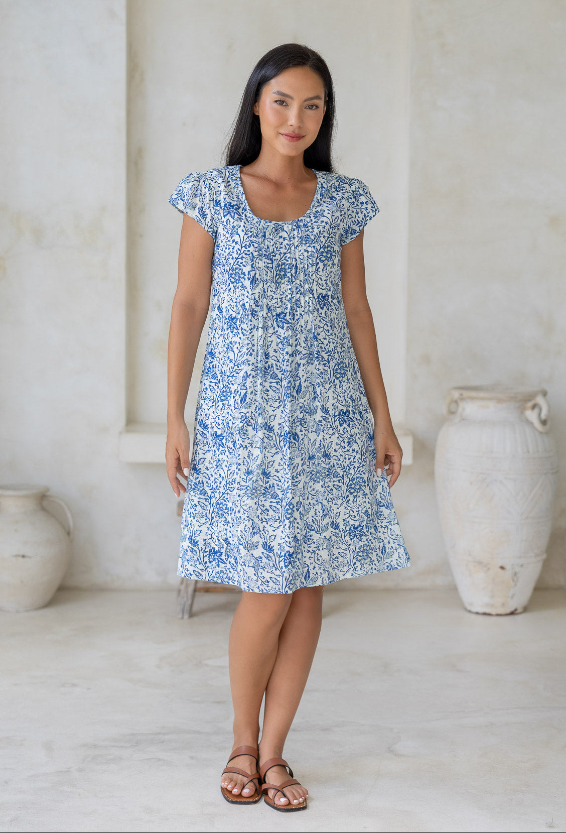 Woman wearing a cap sleeved, above knee length, white based cotton dress with blue floral print and lemon yellow highlights.
