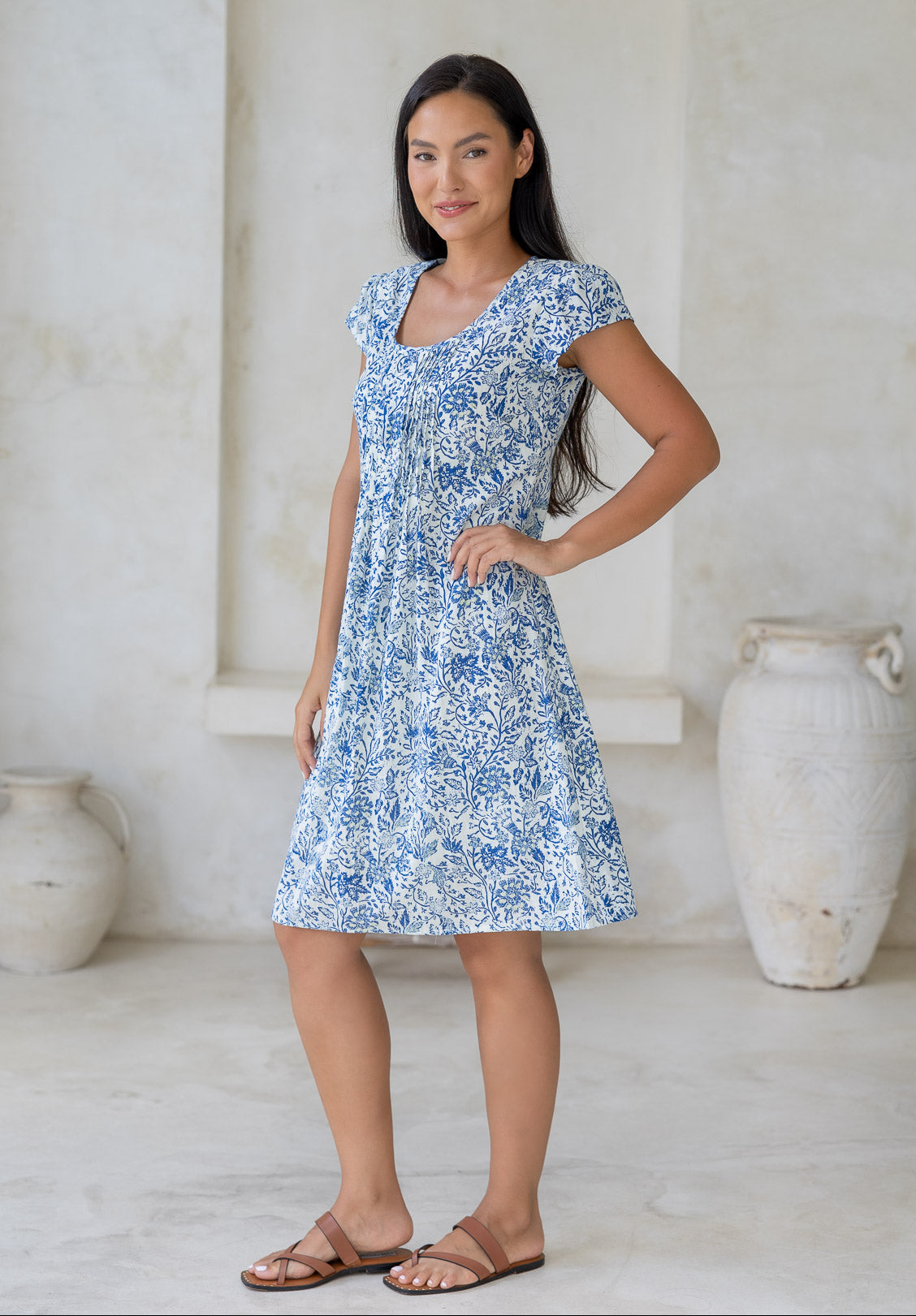 Woman posing in 100% cotton above knee length dress with round neckline, cap sleeves and small pleats across the bodice in white based floral print.