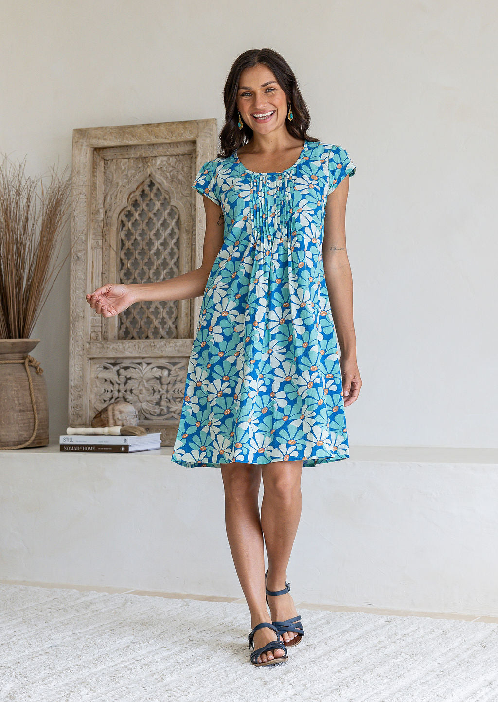 Woman wearing cap sleeved cotton above knee dress in blue and aqua daisy print, with round neckline and pin-tucks across bodice