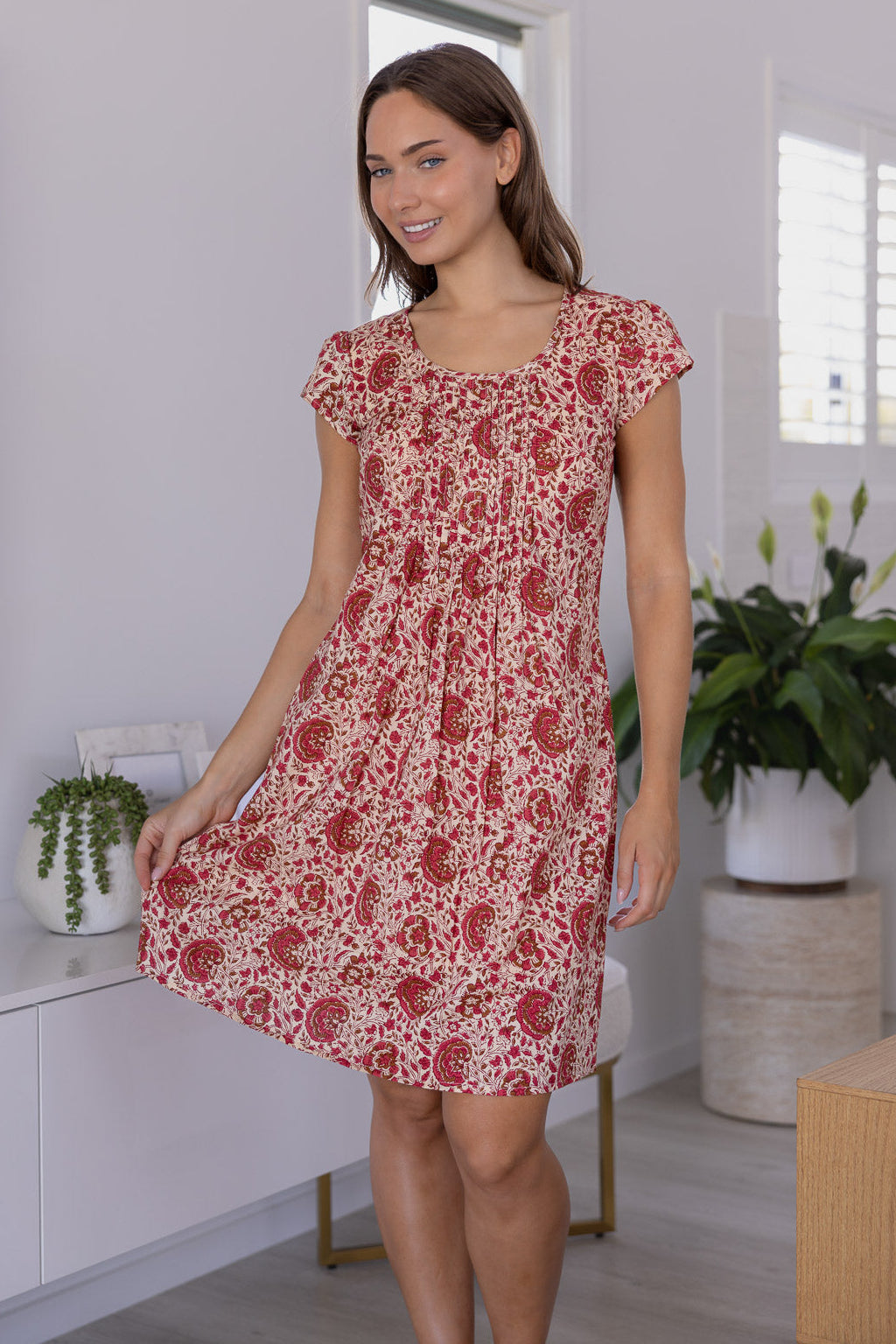 Woman wearing a 100% cotton gentle cream based floral dress in a modern room with white walls and plants.