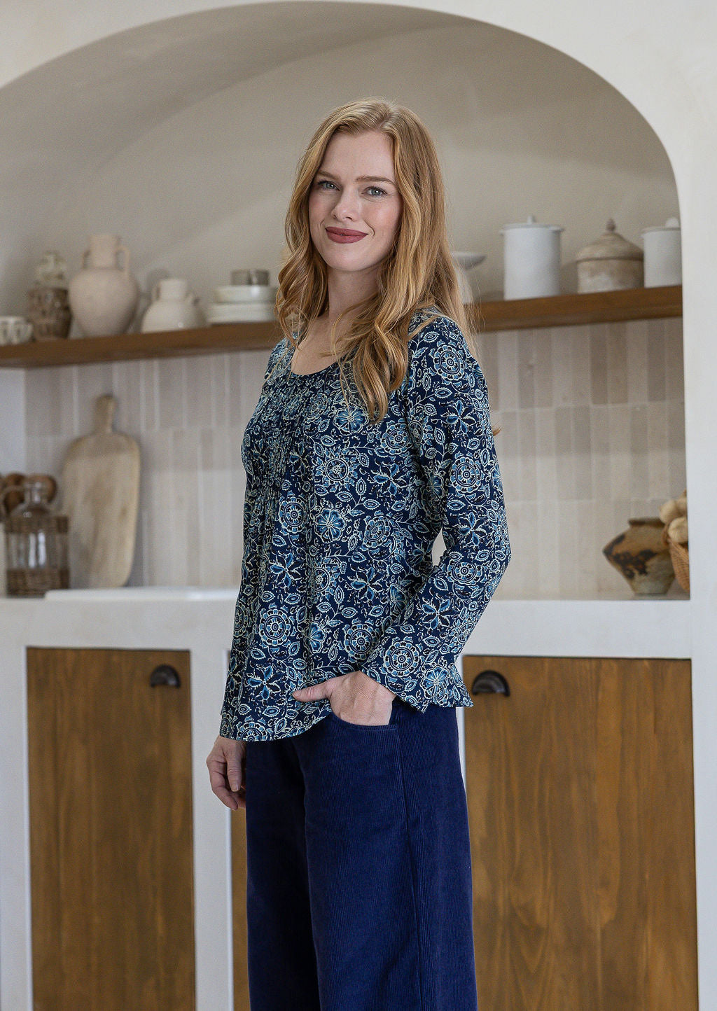 Woman in a blue floral top standing in a kitchen with wooden shelves and white cabinets.