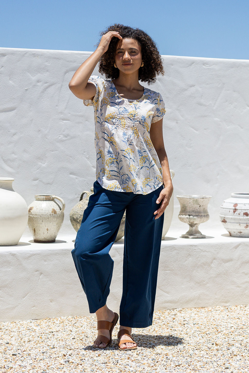 Woman wears neutral based floral print 100% cotton cap sleeve top with pin tuck detail across the bust and a scoop neckline