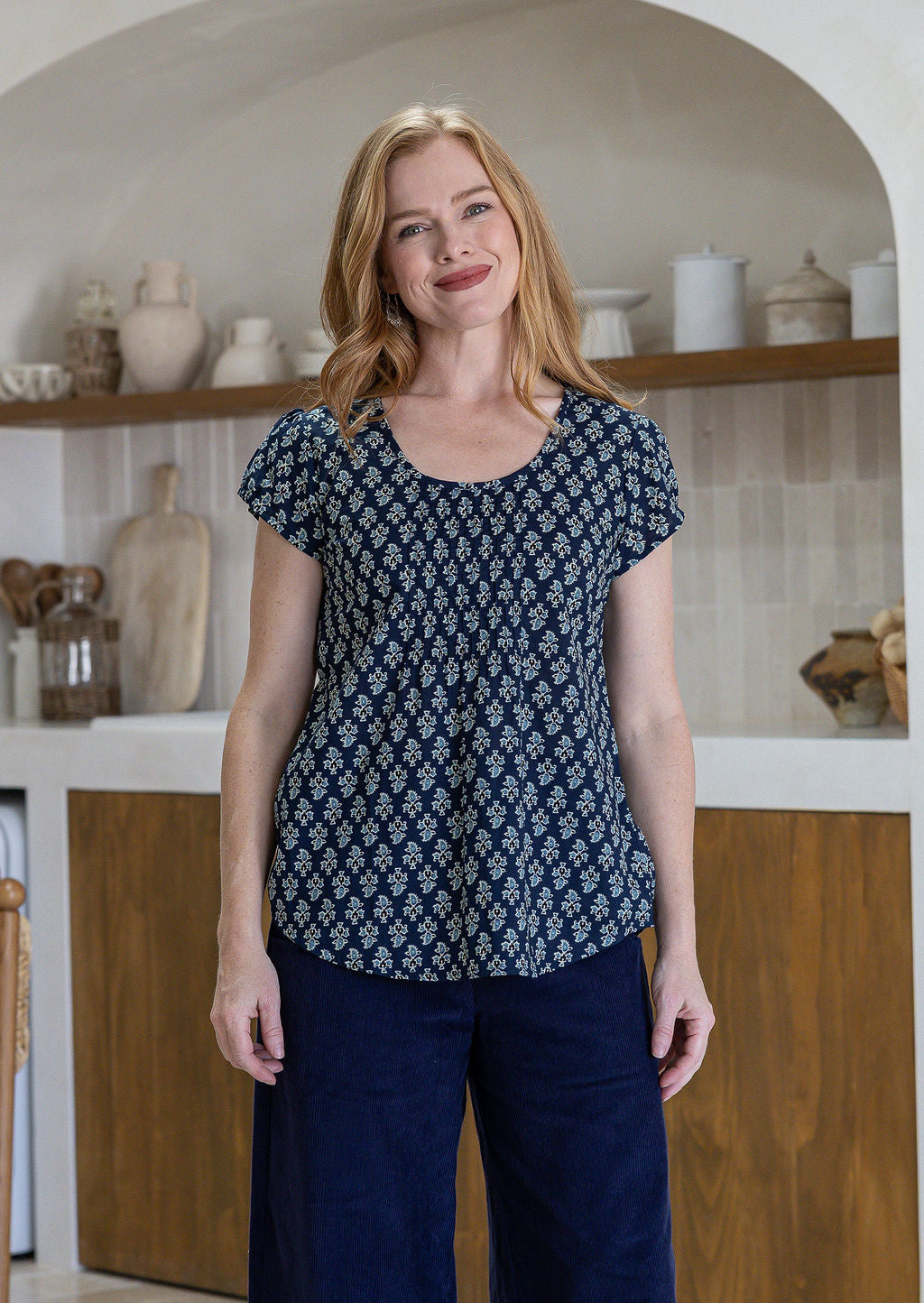 Woman wears navy blue based floral print 100% cotton cap sleeve top with pin tuck detail across the bust and a scoop neckline
