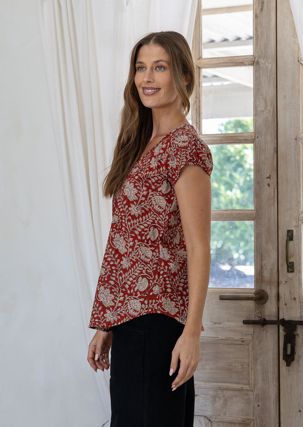 Woman stands sideways wearing 100% cotton top in red based floral print.