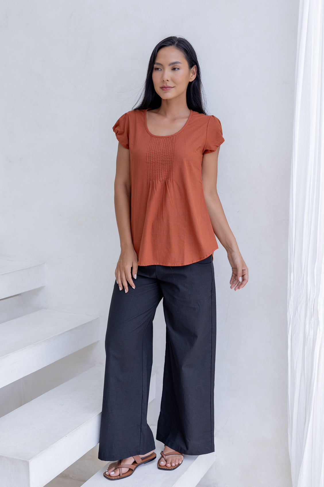 Woman wearing a rust-colored cotton summer top and dark pants standing on a white staircase.