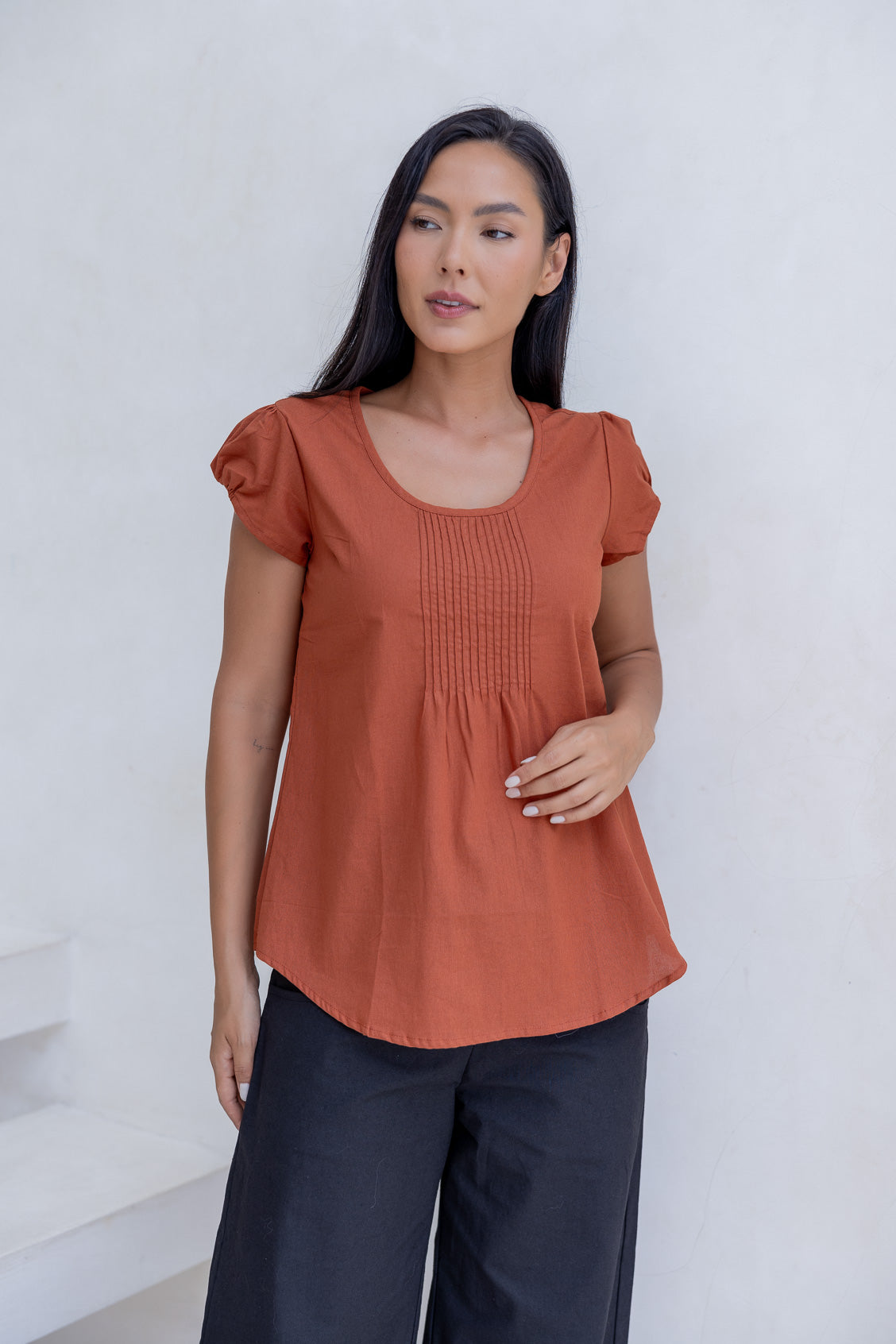 Woman wears 100% cotton cap sleeve top in rust orange shade with pin tuck detail across the bust and a scoop neckline.