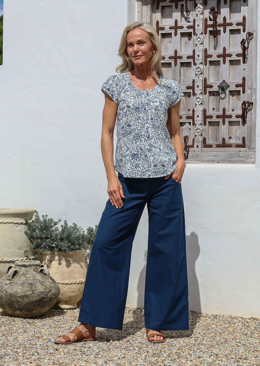 Woman standing with hand in pocket wears 100% cotton short sleeved summer top in white based blue floral print with round neckline and decorative small pleats across the bust.
