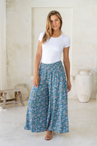Woman wearing a cerulean blue based block style floral print pants with smocked elastic waistband.
