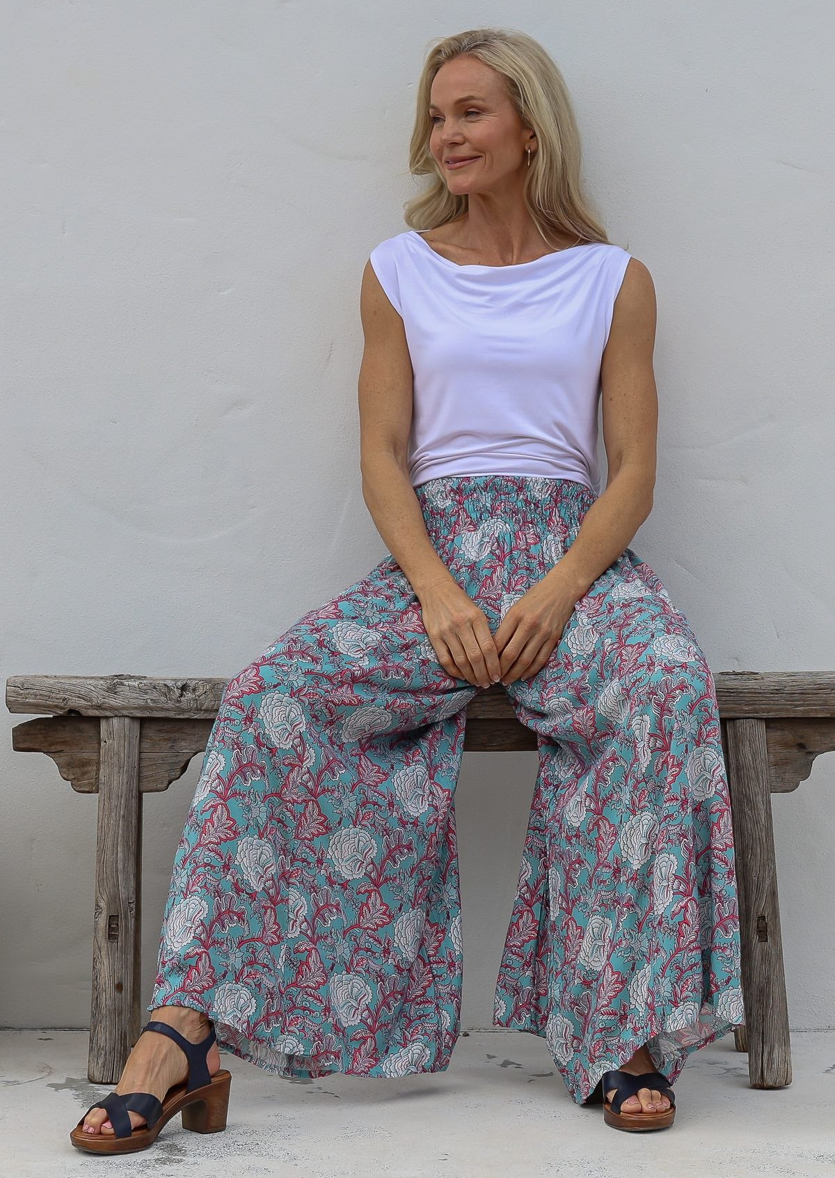 Woman sitting on a wooden bench wearing a white top and soft blue based floral wide leg pants with elastic waist.