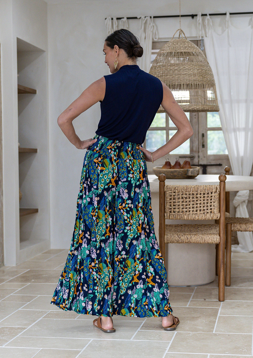 A woman shown from the back wearing a sleeveless navy top and ultra wide rayon pants with a wide shirred waistband in a multicolour floral and peacock print