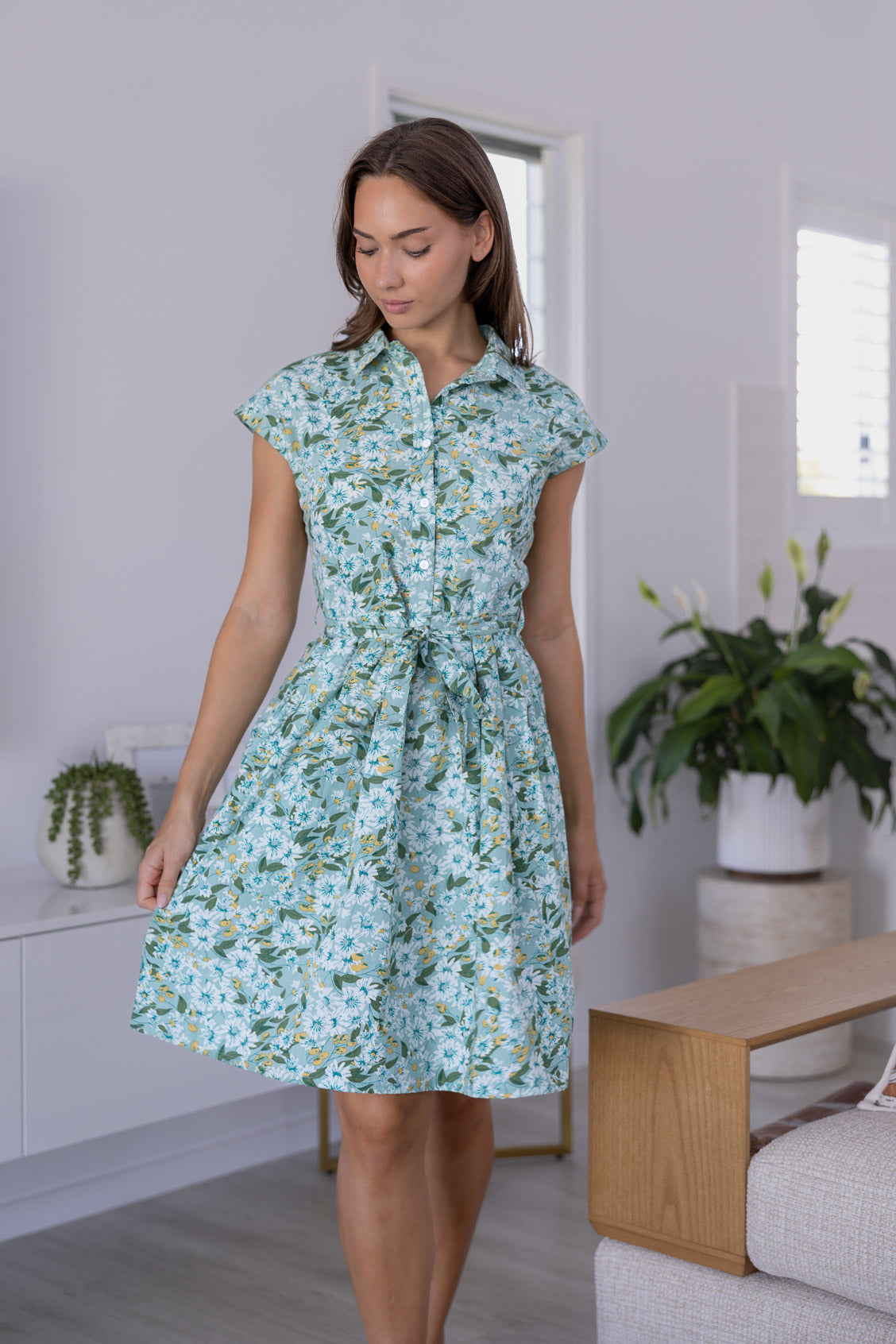 Woman in a sage green floral cotton dress with cap sleeves, buttoned bodice, pockets, box pleats at the waist and a fabric waist tie.