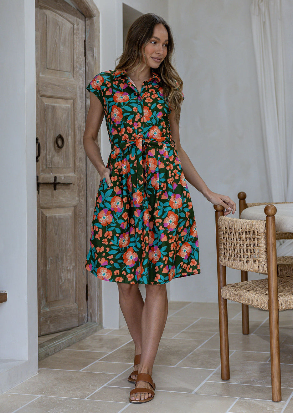Model stands with one hand in pocket wearing a 100% cotton knee length shirt style retro floral dress in olive green based floral print.