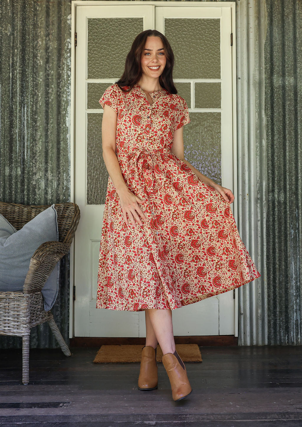 Woman in a floral dress standing in front of a white door with a wicker chair and cushions.