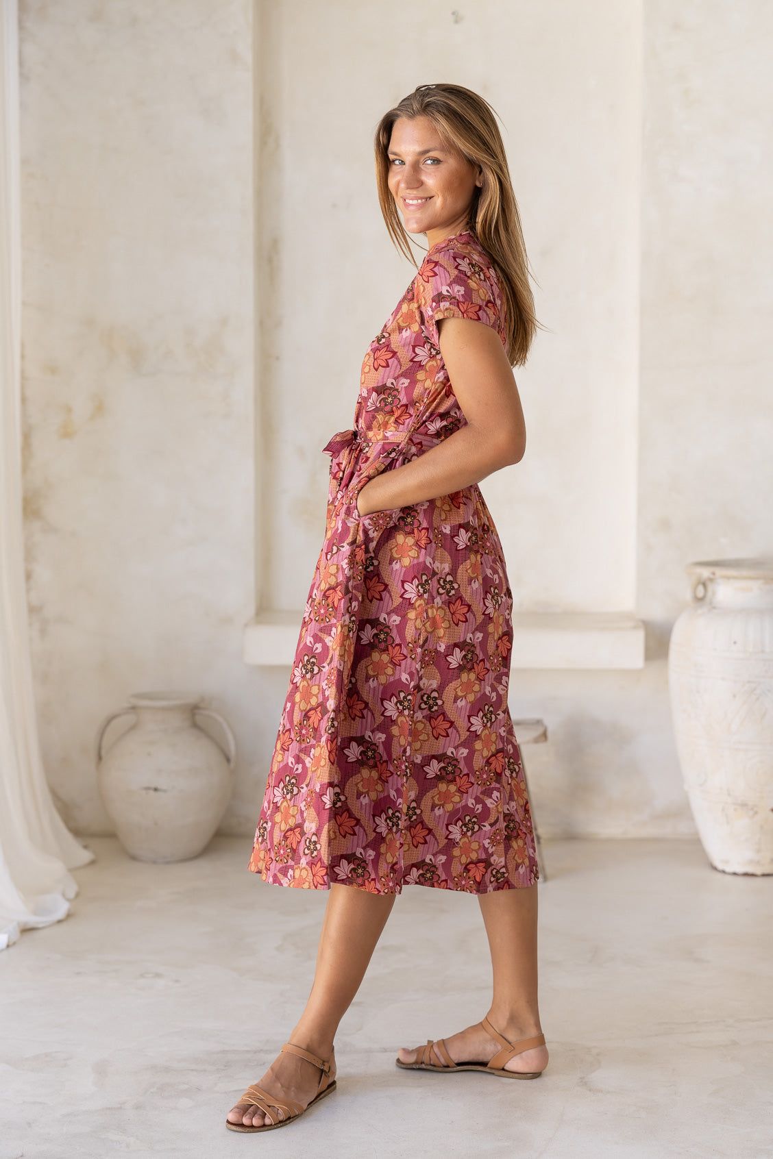 Women stands in side pose with hands in pockets wearing floral printed midi length dress with high round neckline, short sleeves and pockets.
