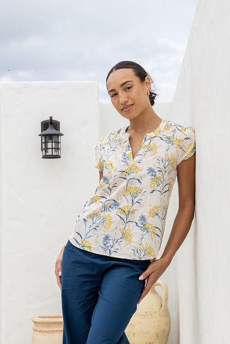 Woman wearing a floral top and blue pants standing against a white wall.