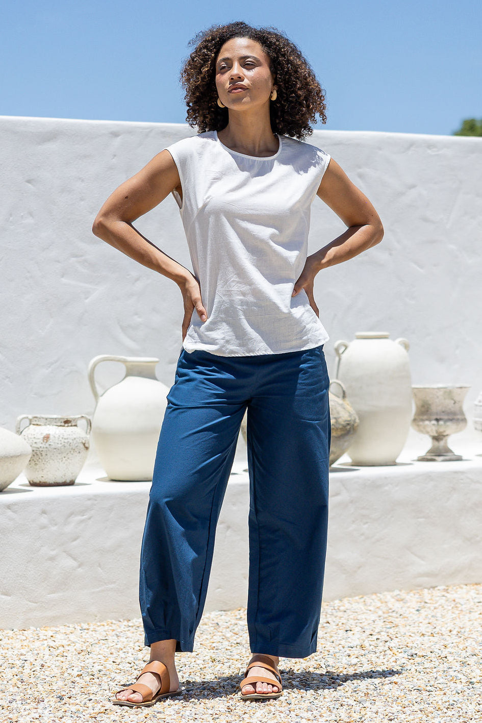 Woman wearing a white cotton sleeveless top with a round neckline, paired with blue pants, with hands on hips
