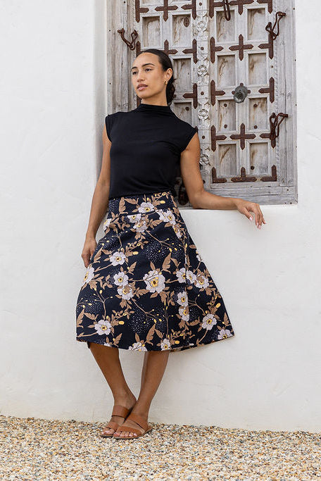 Woman wearing a black top and floral skirt standing against a white wall with a decorative metal door.