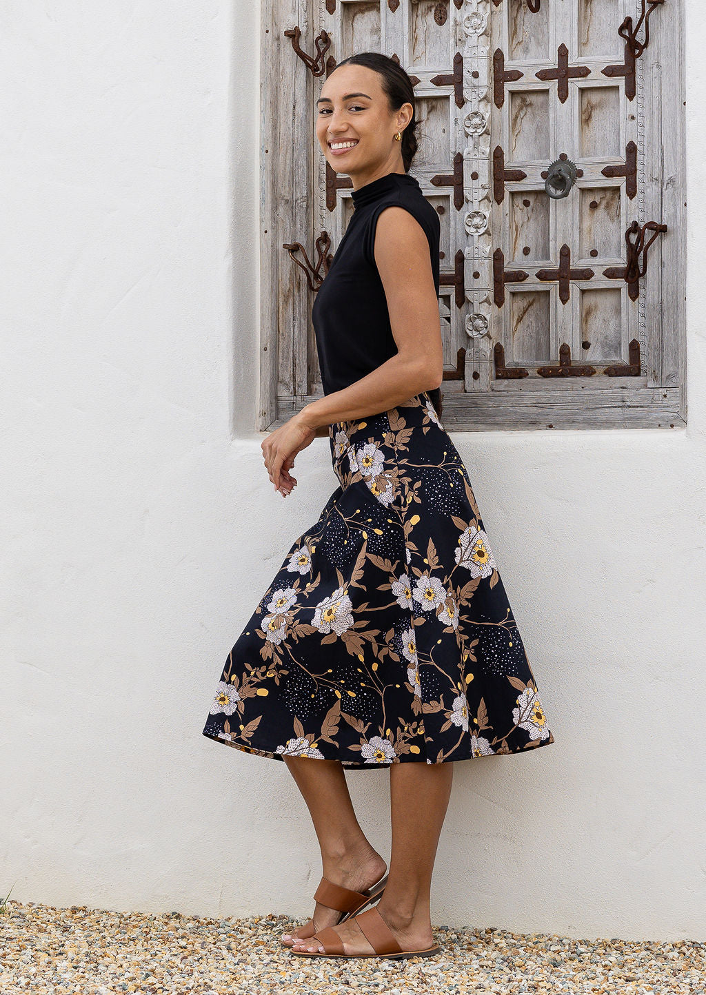 Woman standing sideways wearing a black top and black floral cotton skirt against a wooden window,