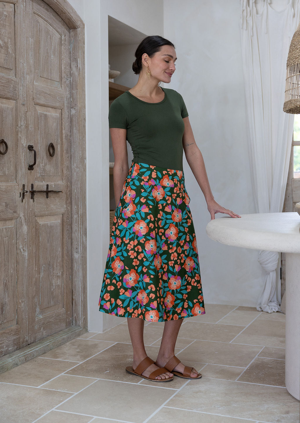 Woman standing sideways wearing a basic top and olive green based floral cotton skirt in a home setting