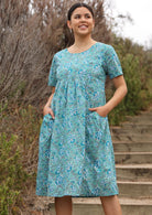 Model wears floral printed cotton dress in aqua shade with short sleeves and hands in pocket.