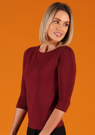 Woman wearing a rayon boat neck maroon 3/4 sleeve top in front of orange background.