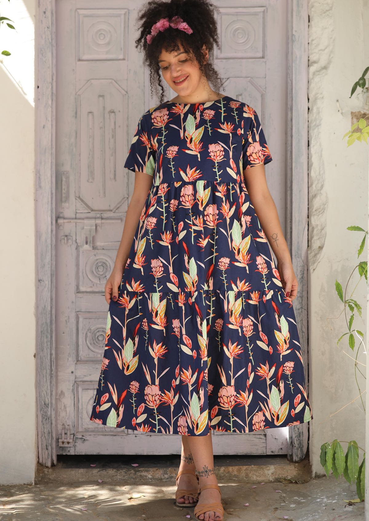 Woman standing in front of white wooden door wears 100% cotton tiered midlength dress in pink and green native floral print.
