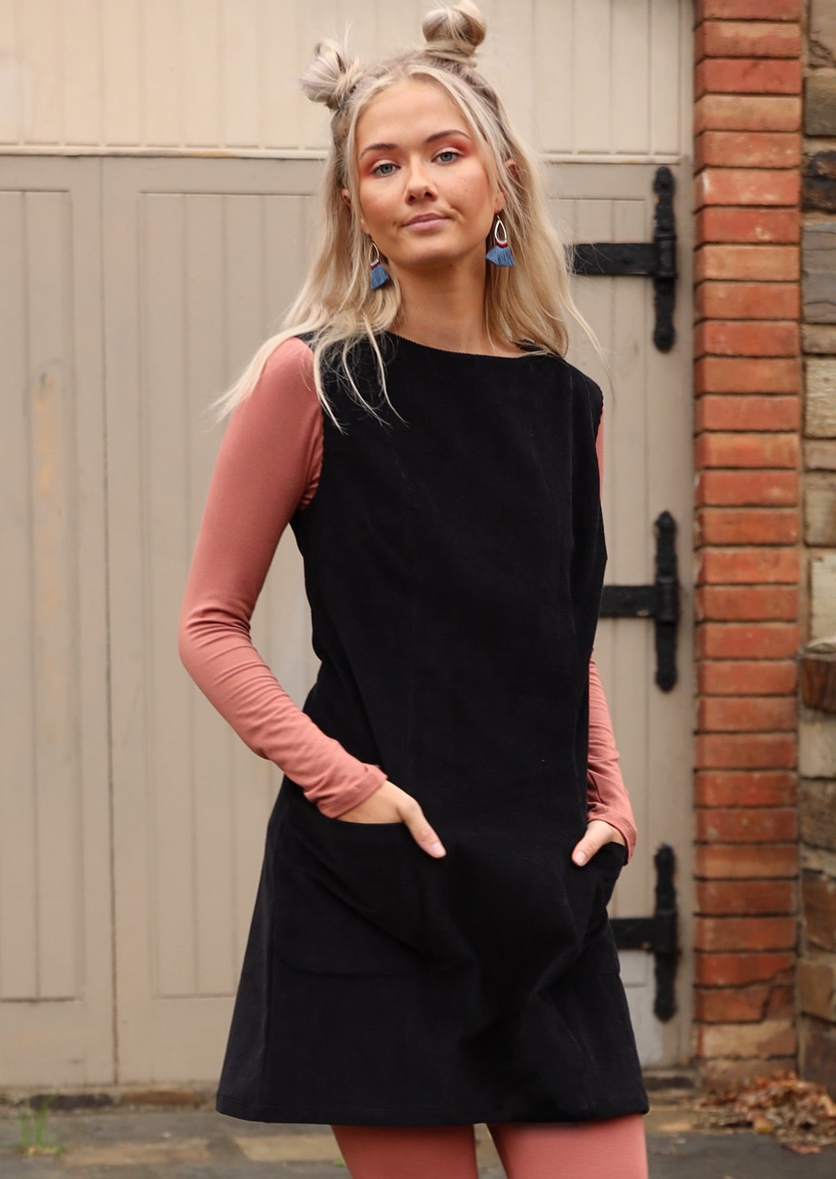 Woman wears black sleeveless cotton corduroy tunic with hands in front pockets