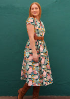 Model standing in front of dark teal background wears colourful retro style dress with boots and stylish belt at the waist.