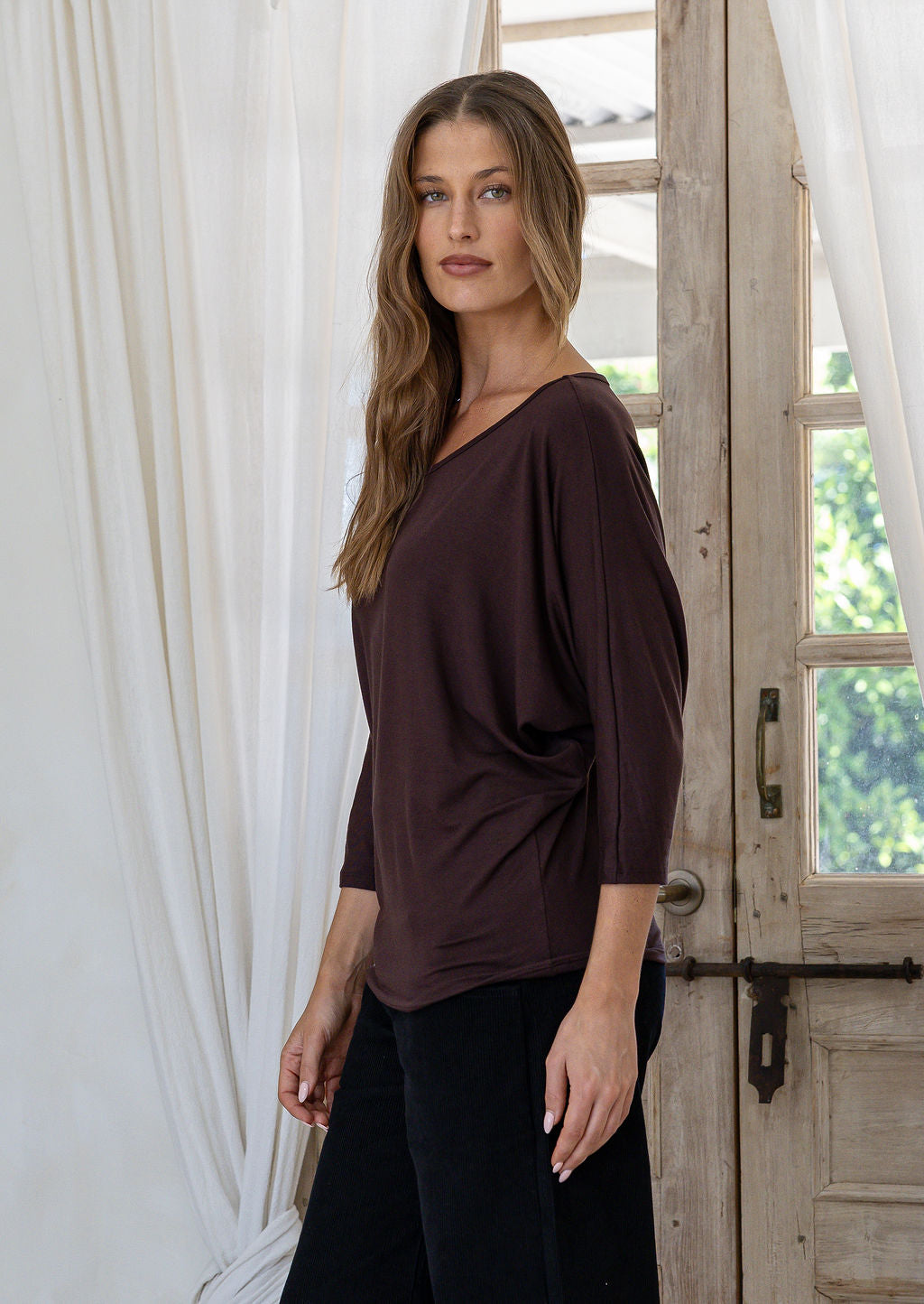 Woman standing side on wearing a brown rayon jersey batwing top with 3/4 sleeves and a round neckline, paired with black pants