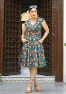 Woman stands in, green floral printed cotton dress with cap sleeves, crossover bodice forming V neckline and hands in pocket.