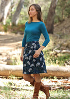Model poses in an A-line corduroy skirt with floral print on dark blue base, paired with a teal top and boots