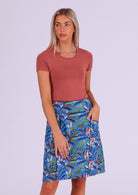 Woman wears cotton A-line above knee skirt in leafy print on a blue base, with hand in pocket 