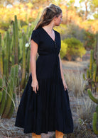 Woman stands in black rayon maxi dress with tulip sleeves, V-neckline and buttoned bodice